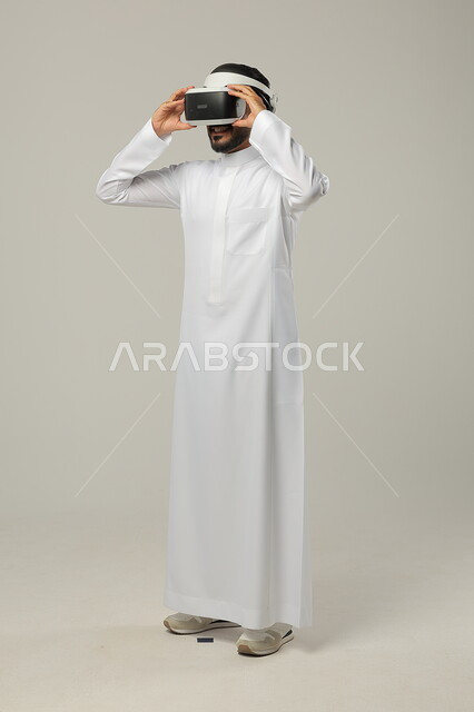 محاكاة العالم الخيالي، بورتريه لرجل عربي خليجي سعودي يرتدي الثوب التقليدي ونظارات الواقع الافتراضي ثلاثية الأبعاد، دمج التقنيات والتكنولوجيا الحديثة مع الترفيه والتسلية، ايماءات الاندماج والتركيز، صورة الجسم بالطول الكامل، خلفية رمادية