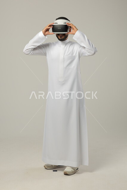 Integrating modern techniques and technology with fun and entertainment, simulating a fantasy world, portrait of a Saudi Gulf Arab man wearing a traditional thobe and 3D virtual reality glasses, gestures of fusion and focus, full-length body portrait, gray background