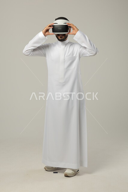 المتعة والتسلية في محاكاة العالم الخيالي، بورتريه لرجل عربي خليجي سعودي يرتدي الثوب التقليدي ويضع نظارات الواقع الافتراضي ثلاثية الأبعاد، استخدام التقنيات والتكنولوجيا الحديثة، ايماءات الاندماج والتركيز، صورة الجسم بالطول الكامل، خلفية رمادية