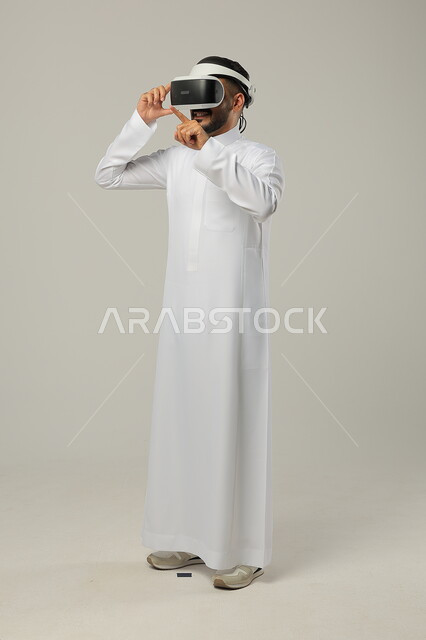 التطور التقني والتكنولوجي، بورتريه لرجل عربي خليجي سعودي يرتدي الثوب التقليدي ونظارات الواقع الافتراضي ثلاثية الأبعاد يحاول النقر والاشارة الى شيء ما، دمج التقنيات والتكنولوجيا الحديثة مع المتعة والتسلية، ايماءات الاندماج والتركيز، صورة الجسم بالطول الكامل، خلفية رمادية