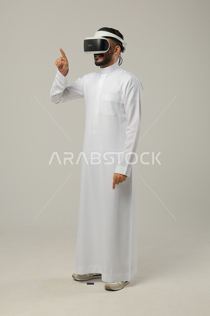 Gestures of integration and focus, simulating a fantasy world, portrait of a Saudi Gulf Arab man wearing a traditional dress and 3D virtual reality glasses trying to click and point at something, merging modern techniques and technology with fun and entertainment, full-length body portrait, gray background