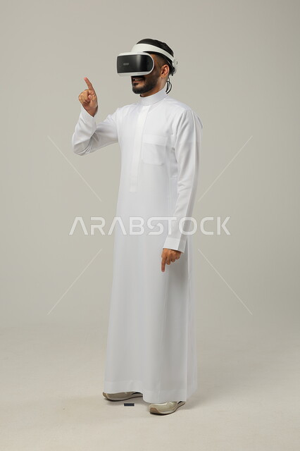 التفاعل ومحاكاة العالم الخيالي، بورتريه لرجل عربي خليجي سعودي يرتدي الثوب التقليدي ونظارات الواقع الافتراضي ثلاثية الأبعاد يحاول النقر والاشارة الى شيء ما، دمج التقنيات والتكنولوجيا الحديثة مع المتعة والتسلية، ايماءات الاندماج والتركيز، صورة الجسم بالطول الكامل، خلفية رمادية