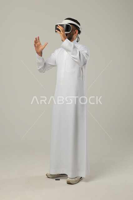 التطور التكنولوجي والتقني، بورتريه لرجل عربي خليجي سعودي يرتدي الثوب التقليدي ونظارات الواقع الافتراضي ثلاثية الأبعاد يحاول تجسيم شيء، دمج التكنواوجياوالتقنيات الحديثة مع المتعة والتسلية، ايماءات الاندماج والتركيز، صورة الجسم بالطول الكامل، خلفية رمادية