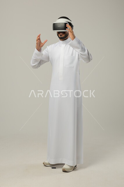 الأجهزة التكنولوجية والتقنيات الحديثة، محاكاة العالم الخيالي، بورتريه لرجل عربي خليجي سعودي يرتدي الثوب التقليدي ونظارات الواقع الافتراضي ثلاثية الأبعاد يحاول تجسيم شيء، ايماءات الاندماج والتركيز، صورة الجسم بالطول الكامل، خلفية رمادية