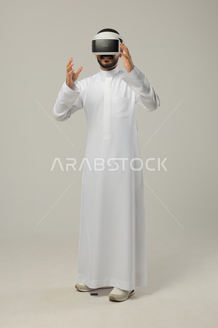 Integrating modern techniques and technology with fun and entertainment, gestures of integration and concentration, simulating the imaginary world, portrait of a Saudi Gulf Arab man wearing a traditional thobe and 3D virtual reality glasses trying to embody something, full-length body image, gray background