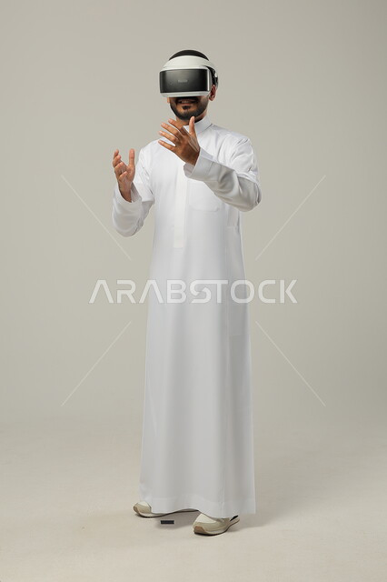 استخدام التقنيات والتكنولوجيا الحديثةوالدمج مع المتعة والتسلية، محاكاة العالم الخيالي، بورتريه لرجل عربي خليجي سعودي يرتدي الثوب التقليدي ونظارات الواقع الافتراضي ثلاثية الأبعاد يحاول تجسيم شيء، ايماءات الاندماج والتركيز، صورة الجسم بالطول الكامل، خلفية رمادية