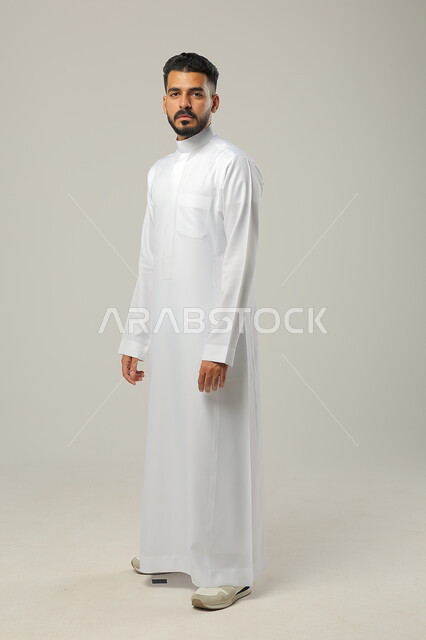 Concern with external appearance and elegance, the concept of masculinity and self-confidence, a portrait of a Saudi Gulf Arab man wearing a traditional dress, looking at the camera with an expression of pleasure, standing upright and firm, full-length body portrait, gray background.