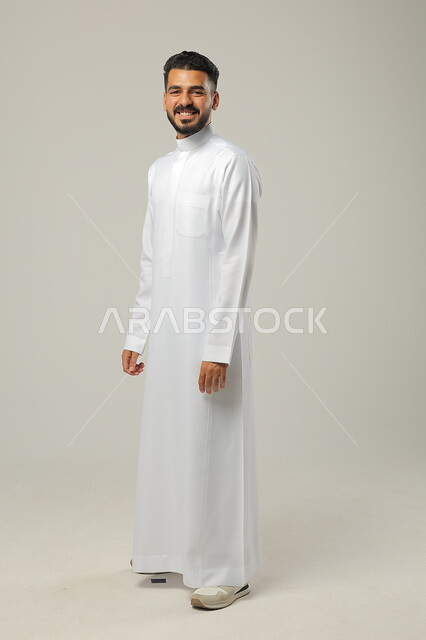  الوقوف بثقة واستقامة، الاهتمام بالمظهر الخارجي والأناقة، مفهوم الرجولة والثقة بالنفس، بورتريه لرجل عربي خليجي سعودي مبتسم  يرتدي الثوب التقليدي ينظر الى الكاميرا، الوقوف باستقامة وثبات، صورة الجسم بالطول الكامل، خلفية رمادية