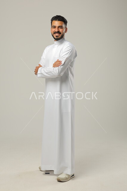 تعابير السرور والبهجة، الاهتمام بالمظهر الخارجي والأناقة، مفهوم الرجولة والثقة بالنفس، بورتريه لرجل عربي خليجي سعودي مبتسم يرتدي الثوب التقليدي ينظر الى الكاميرا، الوقوف باستقامة وثبات بايدي متقاطعة، صورة الجسم بالطول الكامل، خلفية رمادية