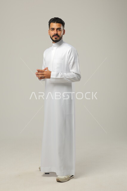 تعابير السعادة والسرور، بورتريه لرجل عربي خليجي سعودي يرتدي الثوب التقليدي يرفع يديه بحركة تلامس الأصابع، مفهوم الذكاء والمعرفة، النظر الى الكاميرا يايماءات الثقة بالنفس، صورة الجسم بالطول الكامل، خلفية رمادية