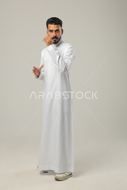 Silence and stopping talking, portrait of a Saudi Gulf Arab young man standing upright, making spontaneous movements and looking at the camera with offensive gestures, full-length body portrait, gray background
