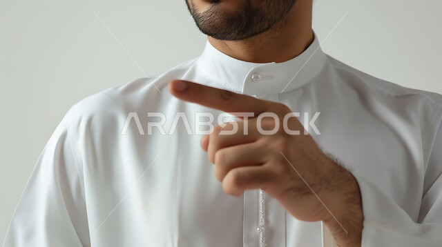 الاشارة نحو شيء وتحديد هدف ما، حركة يد تدل على هذا المكان (هنا)، بورتريه مقرب لرجل عربي خليجي سعودي يرتدي الثوب التقليدي يشير بأصبع السبابة على مكان معين، خلفية بيضاء