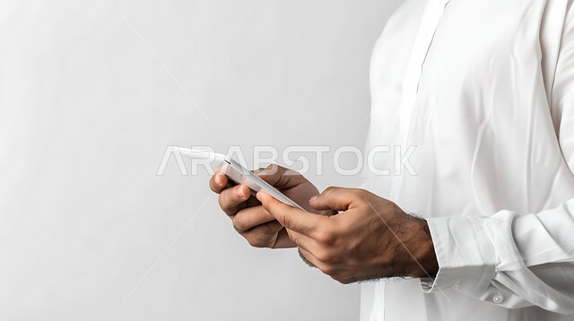 التسلية و الترفيه باستخدام الأجهزة التقنية الحديثة, التواصل مع الأهل و الأصدقاء, بورتريه مقرب لرجل عربي خليجي سعودي يرتدي الثوب التقليدي يمسك بيده الهاتف المحمول، خلفية بيضاء