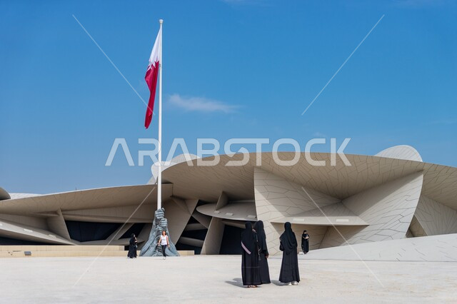Visiting famous tourist cultural heritage landmarks, the flagpole of the State of Qatar in front of the National Museum building in the shape of a desert rose during the day, advanced architectural engineering art for museums, artistic decorations that reflect the Qatari culture and the country’s identity.