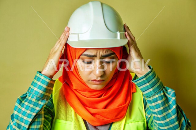 عمل النساء في القطاع الهندسي في المملكة العربية السعودية، بورتريه مقرب لفتاة عربية خليجية سعودية محجبة ترتدي الخوذة وسترة الحماية وتمسك رأسها بيديها بإيماءات الألم، الشعور بالتعب والإرهاق والحاجة إلى الراحة، مهندسة تعاني من ضغط العمل، خلفية ملونة