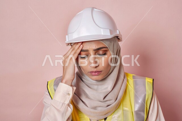  الشعور بالتعب والإرهاق والحاجة إلى الراحة، عمل النساء في القطاع الهندسي في المملكة العربية السعودية، بورتريه مقرب لفتاة عربية خليجية سعودية محجبة ترتدي الخوذة وسترة الحماية وتمسك رأسها بيديها بإيماءات الألم، مهندسة تعاني من ضغط العمل، خلفية ملونة