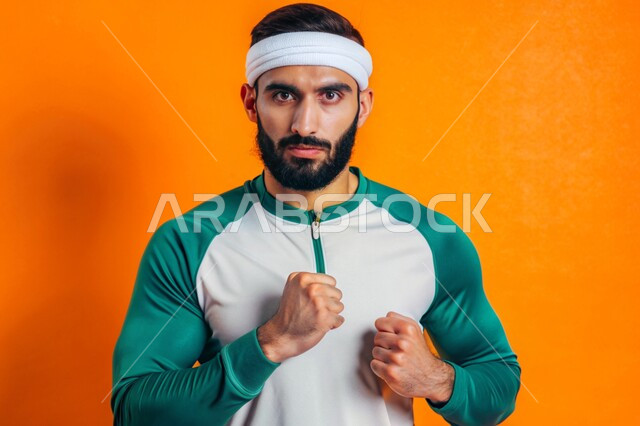 التمتع بالبنية الجسدية القوية، بورتريه لشاب عربي خليجي سعودي يرتدي الزي الرياضي ينظر الى الكاميرا بإيماءات الثقة بالنفس يقوم بممارسة التمارين الرياضية، مفهوم القوة العضلية، اللياقة البدنية والجسم الصحي المتين، خلفية ملونة