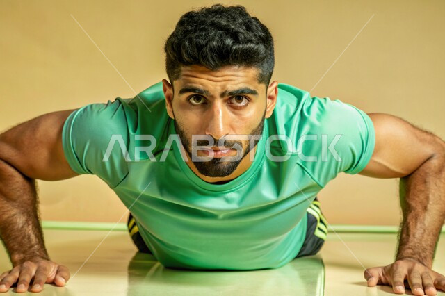 التمتع بالبنية الجسدية القوية، بورتريه لشاب عربي خليجي سعودي يرتدي الزي الرياضي ينظر الى الكاميرا بإيماءات الثقة بالنفس يقوم بممارسة تمارين الضغط، مفهوم القوة العضلية، اللياقة البدنية والجسم الصحي المتين، خلفية بيج