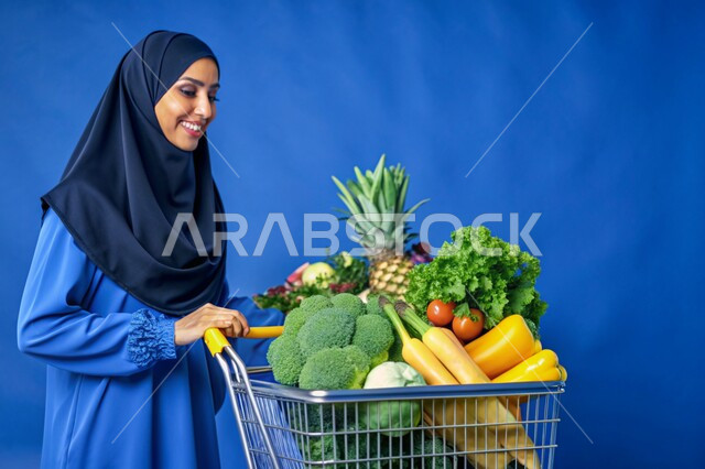 محاصيل زراعية ومنتجات محلية وطنية، بوتريه مقرب لسيده عربيه خليجيه سعوديه محجبة مبتسمة تمسك بيدها عربة التسوق بإيماءات الفرح والسرور، اختيار أجود أنواع الخضار والفواكه الطازجة، خلفية زرقاء