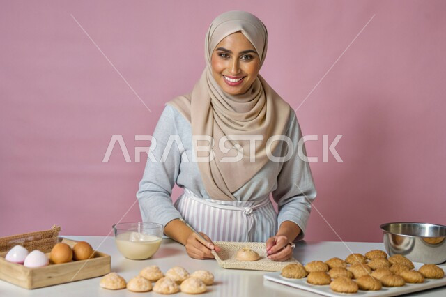 Preparing desserts with fresh natural ingredients, cooking skills and arts, close-up portrait of a veiled Saudi Arabian Gulf woman, smiling, looking at the camera, standing inside the kitchen preparing food recipes with gestures of pleasure and happiness, pink background