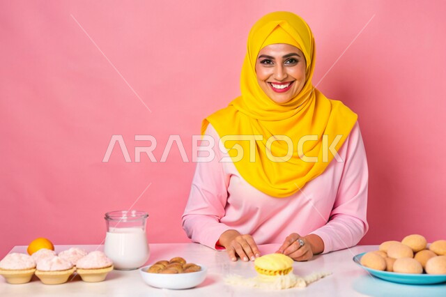 إعداد الحلويات بمكونات طبيعية طازجة، مهارات وفنون الطبخ, بورتريه مقرب لإمرأة عربية خليجية سعودية محجبة مبتسمة تنظر الى الكاميرا تقف داخل المطبخ تقوم بتحضير وصفات الطعام بإيماءات السرور والسعادة، خلفية وردية