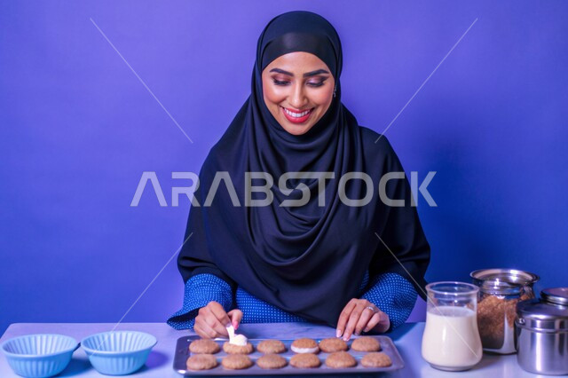 مهارات وفنون الطبخ, إعداد الحلويات بمكونات طبيعية طازجة، بورتريه مقرب لإمرأة عربية خليجية سعودية محجبة مبتسمة تقوم بتحضير وصفات الطعام بإيماءات السرور والسعادة، خلفية زرقاء