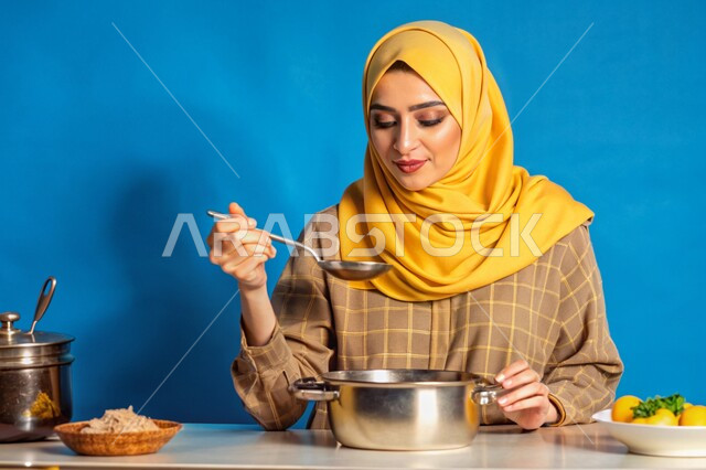 مهارات وفنون الطبخ, إعداد الأطعمة بمكونات طبيعية طازجة، بورتريه مقرب لإمرأة عربية خليجية سعودية محجبة مبتسمة تقوم بتذوق النكهات بإيماءات السرور والسعادة، تحضير وصفات الطعام، خلفية زرقاء