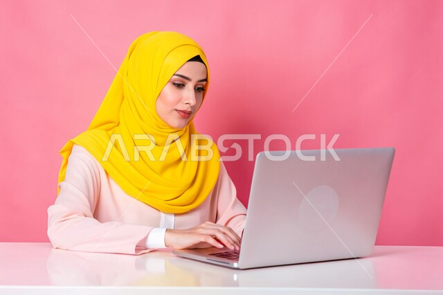 مفهوم التعليم و العمل عن بعد، بورتريه مقرب لشابة عربية خليجية سعودية محجبة مبتسمة تعمل على جهاز الحاسوب المحمول، إستخدام الأجهزة التقنية الحديثة، التسويق الإلكتروني وتصفح الإنترنت، مشاهدة الأفلام عبر اللابتوب، مهن ووظائف نسائية، خلفية وردية