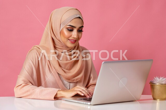 مفهوم التعليم و العمل عن بعد، بورتريه مقرب لشابة عربية خليجية سعودية محجبة مبتسمة تعمل على جهاز الحاسوب المحمول، إستخدام الأجهزة التقنية الحديثة، التسويق الإلكتروني وتصفح الإنترنت، مشاهدة الأفلام عبر اللابتوب، مهن ووظائف نسائية، خلفية وردية