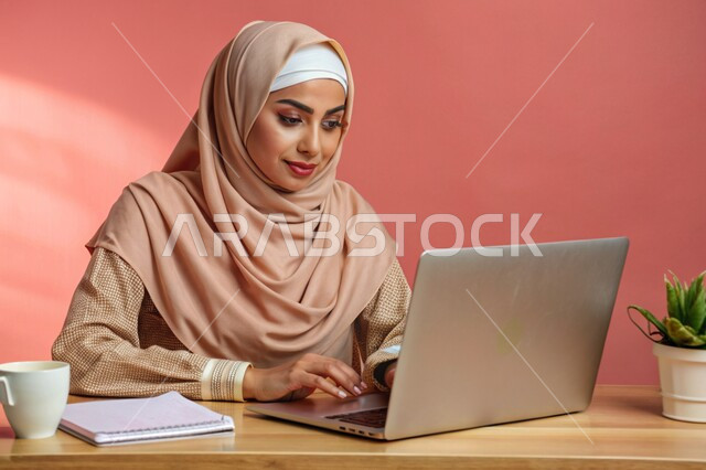 مفهوم التعليم و العمل عن بعد، بورتريه مقرب لشابة عربية خليجية سعودية محجبة مبتسمة تعمل على جهاز الحاسوب المحمول، إستخدام الأجهزة التقنية الحديثة، التسويق الإلكتروني وتصفح الإنترنت، مشاهدة الأفلام عبر اللابتوب، مهن ووظائف نسائية، خلفية وردية