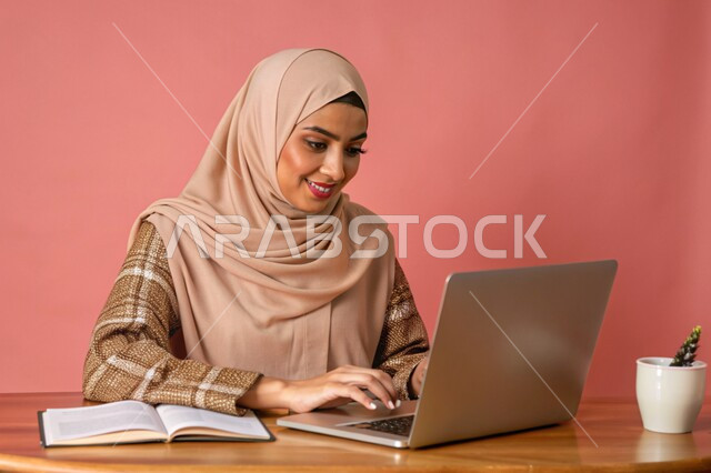 مفهوم التعليم و العمل عن بعد، بورتريه مقرب لشابة عربية خليجية سعودية محجبة مبتسمة تعمل على جهاز الحاسوب المحمول، إستخدام الأجهزة التقنية الحديثة، التسويق الإلكتروني وتصفح الإنترنت، مشاهدة الأفلام عبر اللابتوب، مهن ووظائف نسائية، خلفية وردية