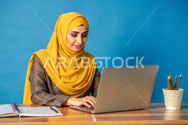 مفهوم التعليم و العمل عن بعد، بورتريه مقرب لشابة عربية خليجية سعودية محجبة مبتسمة تعمل على جهاز الحاسوب المحمول، إستخدام الأجهزة التقنية الحديثة، التسويق الإلكتروني وتصفح الإنترنت، مشاهدة الأفلام عبر اللابتوب، مهن ووظائف نسائية، خلفية زرقاء