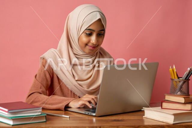 إستخدام الأجهزة التقنية الحديثة، مفهوم التعليم و العمل عن بعد، بورتريه مقرب لفتاة عربية خليجية سعودية محجبة مبتسمة تعمل على جهاز الحاسوب المحمول، جودة التعليم وتطور الوسائل التعليمية في المملكة العربية السعودية، خلفية وردية