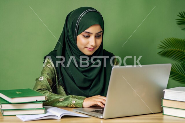 جودة التعليم وتطور الوسائل التعليمية في المملكة العربية السعودية، إستخدام الأجهزة التقنية الحديثة، مفهوم التعليم و العمل عن بعد، بورتريه مقرب لفتاة عربية خليجية سعودية محجبة مبتسمة تعمل على جهاز الحاسوب المحمول، خلفية خضراء
