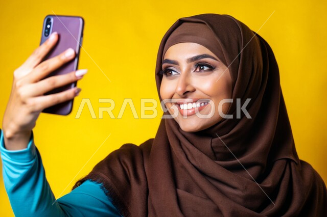 Documenting happy moments, using modern technical devices, a close-up portrait of a veiled Saudi Arabian Gulf woman, smiling, holding a mobile phone in her hand, taking souvenir selfies, various mobile programs and applications, yellow background