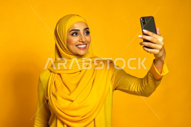 توثيق اللحظات السعيدة، إستخدام الأجهزة التقنية الحديثة، بورتريه مقرب لشابة عربية خليجية سعودية محجبة مبتسمة تمسك بيدها جهاز الهاتف المحمول تقوم بالتقاط صور السيلفي التذكارية، برامج وتطبيقات الجوال المتنوعة، خلفية صفراء