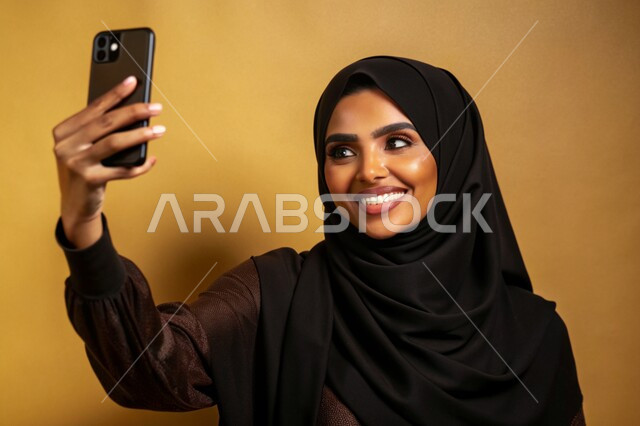 توثيق اللحظات السعيدة، إستخدام الأجهزة التقنية الحديثة، بورتريه مقرب لشابة عربية خليجية سعودية محجبة مبتسمة تمسك بيدها جهاز الهاتف المحمول تقوم بالتقاط صور السيلفي التذكارية، برامج وتطبيقات الجوال المتنوعة، خلفية ملونة