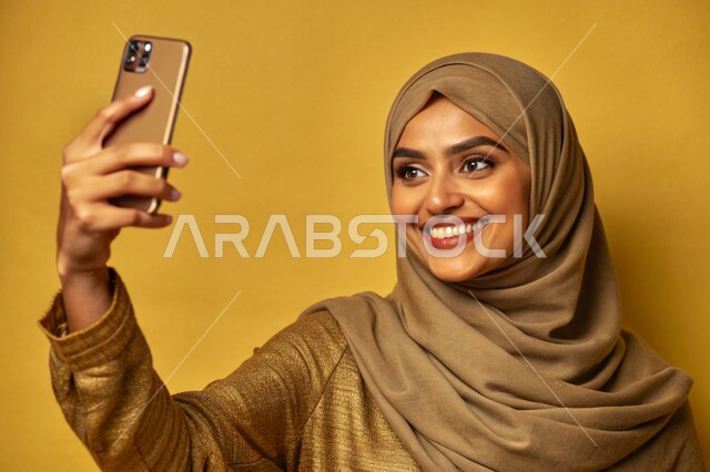 توثيق اللحظات السعيدة، إستخدام الأجهزة التقنية الحديثة، بورتريه مقرب لشابة عربية خليجية سعودية محجبة مبتسمة تمسك بيدها جهاز الهاتف المحمول تقوم بالتقاط صور السيلفي التذكارية، برامج وتطبيقات الجوال المتنوعة، خلفية ملونة