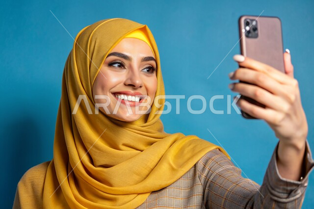 Various mobile programs and applications, documenting happy moments, using modern technical devices, a close-up portrait of a veiled Saudi Arabian Gulf woman, smiling, holding a mobile phone in her hand, taking souvenir selfies, blue background