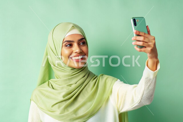 برامج وتطبيقات الجوال المتنوعة، توثيق اللحظات السعيدة، إستخدام الأجهزة التقنية الحديثة، بورتريه مقرب لشابة عربية خليجية سعودية محجبة مبتسمة تمسك بيدها جهاز الهاتف المحمول تقوم بالتقاط صور السيلفي التذكارية، خلفية خضراء