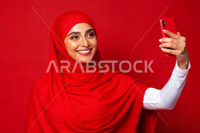 برامج وتطبيقات الجوال المتنوعة، توثيق اللحظات السعيدة، إستخدام الأجهزة التقنية الحديثة، بورتريه مقرب لشابة عربية خليجية سعودية محجبة مبتسمة تمسك بيدها جهاز الهاتف المحمول تقوم بالتقاط صور السيلفي التذكارية، خلفية حمراء