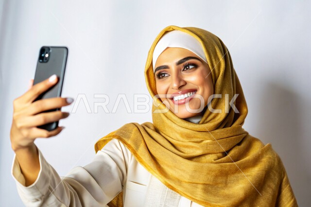 برامج وتطبيقات الجوال المتنوعة، توثيق اللحظات السعيدة، إستخدام الأجهزة التقنية الحديثة، بورتريه مقرب لشابة عربية خليجية سعودية محجبة مبتسمة تمسك بيدها جهاز الهاتف المحمول تقوم بالتقاط صور السيلفي التذكارية، خلفية بيضاء