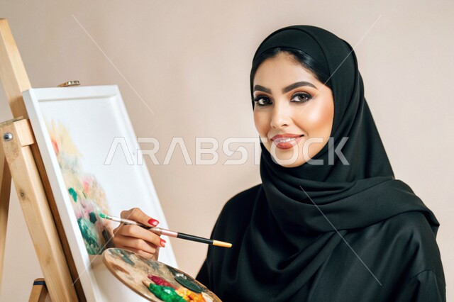 نشاطات ترفيهية فنية، قضاء اوقات ممتعة في قاعة المرسم, بورتريه مقرب لشابة عربية خليجية سعودية محجبة تحمل بيدها بالتة ألوان تنظر الى الكاميرا تقوم بالرسم على اللوحة, معرض الرسوم التشكيلية، تنمية المهارات والفنون اليدوية, خلفية بيضاء