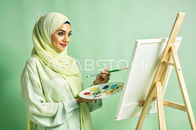 معرض الرسوم التشكيلية، نشاطات ترفيهية فنية، قضاء اوقات ممتعة في قاعة المرسم, بورتريه مقرب لشابة عربية خليجية سعودية محجبة تحمل بيدها بالتة ألوان وتقوم بالرسم على اللوحة, تنمية المهارات والفنون اليدوية, خلفية خضراء