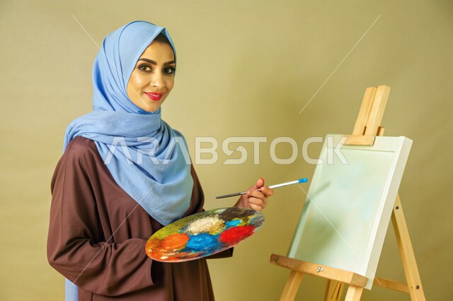 معرض الرسوم التشكيلية، نشاطات ترفيهية فنية، قضاء اوقات ممتعة في قاعة المرسم, بورتريه مقرب لشابة عربية خليجية سعودية محجبة تنظر الى الكاميرا تحمل بيدها بالتة ألوان تقوم بالرسم على اللوحة, تنمية المهارات والفنون اليدوية, خلفية ملونة