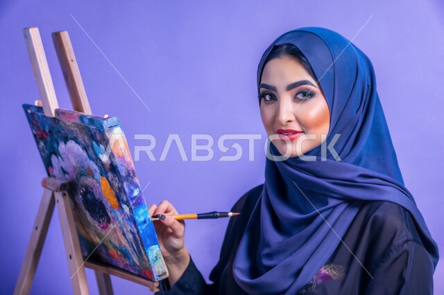 معرض الرسوم التشكيلية، نشاطات ترفيهية فنية، قضاء اوقات ممتعة في قاعة المرسم, بورتريه مقرب لشابة عربية خليجية سعودية محجبة تنظر الى الكاميرا تحمل بيدها بالتة ألوان تقوم بالرسم على اللوحة, تنمية المهارات والفنون اليدوية, خلفية زرقاء