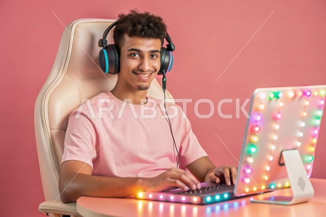 منصة البث المباشر في عالم ألعاب الفيديو، بورتريه لشاب عربي خليجي سعودي يرتدي سماعة الرأس ينظر الى الكاميرا يعمل على جهاز الحاسوب المحمول، استخدام التقنيات والتكنولوجيا الحديثة، مفهوم الاندماج والتفاعل، خلفية وردية
