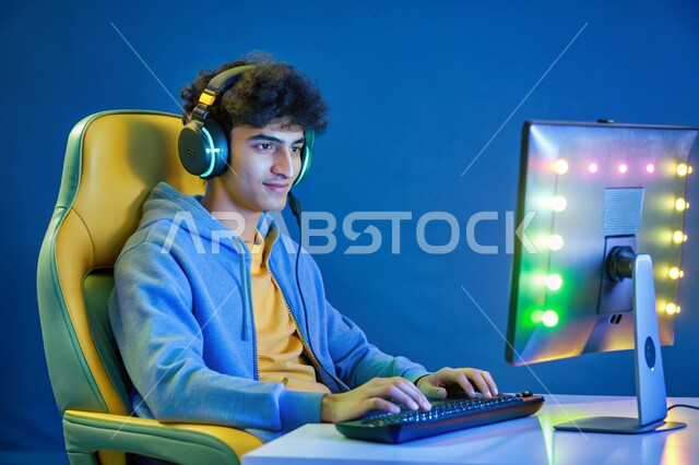 استخدام التقنيات والتكنولوجيا الحديثة، منصة البث المباشر في عالم ألعاب الفيديو، بورتريه لشاب عربي خليجي سعودي يرتدي سماعة الرأس يعمل على جهاز الحاسوب المحمول، مفهوم الاندماج والتفاعل، خلفية زرقاء