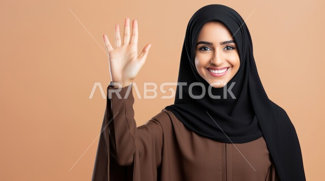 النظر الى الكاميرا بإيماءات السرور، مفهوم الأنوثة والنعومة، الاهتمام بالأناقة والمظهر الخارجي، بورتريه مقرب لامرأة عربية خليجية سعودية محجبة تقف وترفع يدها للنهي عن شيء ما، خلفية بيج