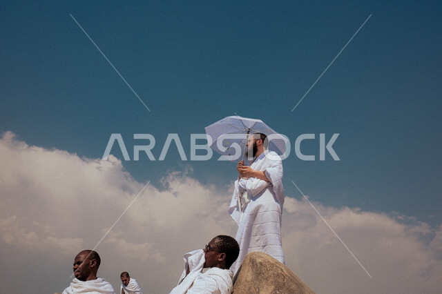 العبادة والتقرب إلى الله بالأعمال الصالحة، شاب خليجي عربي سعودي يرتدي ملابس الاحرام ويحمل بيده مظلة يقف على جبل عرفات، آداء مناسك الحج والعمرة، أماكن ومعالم دينية اسلامية مقدسة في المملكة العربية السعودية